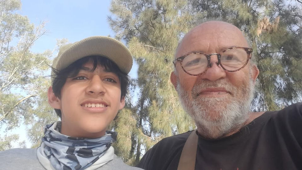 A young person and an older person with glasses smiling outdoors. The background has tall trees and a clear blue sky.
