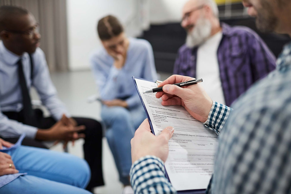 A group in a discussion, seated in a circle. Person in plaid shirt holds a pen and clipboard. The setting is a bright, neutral room.