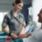 A nurse in a camouflage uniform smiles kindly at a patient in a hospital bed. An IV drip is visible in the background, conveying a caring mood.