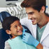 Dentist in a white coat and gloves smiles at a boy in a dental chair. The boy grins, wearing a blue bib. Dental tools in the background.