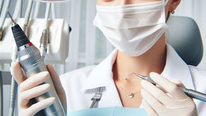 Dentist in white coat and mask holds dental drill and mirror under bright exam light, set in a dental office. Mood is professional.