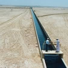 Two people stand on a structure over a long water channel in a desert setting. The scene is sunny, with clear blue sky and barren land.