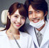 Smiling dentist and a patient in white coats. Dentist holds a mirror tool, gray background. Both show a cheerful, friendly mood.