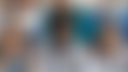 Students in lab coats and scrubs focus on a microscope in a science lab. Test tubes with colorful liquids surround them. Mood is attentive.