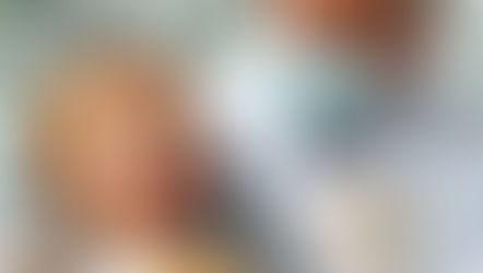 A dentist in a mask and gloves checks a smiling girl's teeth. She holds a teddy bear. Bright dental office setting.