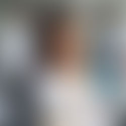 Smiling dentist in white coat and gloves in foreground. Two masked dental assistants work on a patient in a modern clinic. Calm atmosphere.