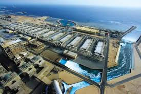 Aerial view of a large desalination plant by the sea, with arranged white structures, blue water channels, and vast ocean in the background.