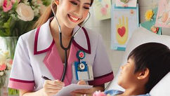 Nurse in white and pink uniform smiles while listening to a young boy's heart with a stethoscope. Flowers and colorful cards in background.