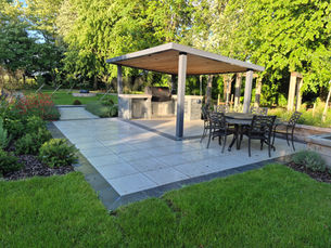 Modern patio with pergola in warm sunlight with an outdoor bbq kitchen , and wrought iron round table with chairs. The patio is surrounded by lush green lawn and planting.