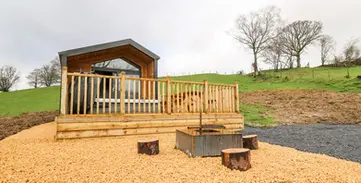 Wooden lodge with deck, fire pit, log seats, green hills beyond.