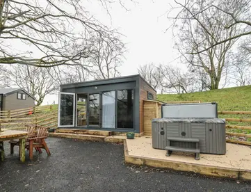 Modern cabin with large windows, outdoor hot tub, and grassy hill.