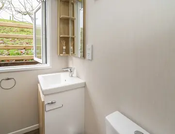 Small bathroom with white sink, toilet, open window showing grassy bank.