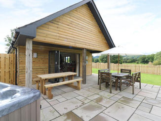 Wooden lodge with patio, outdoor dining area, hot tub, green hills.