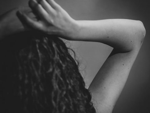 A woman's head seen from the back. Her hands rest on top of her dark curly hair