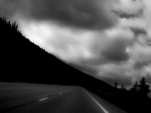 Dark image of a road lined by forest and a car in the distance, with heavy and dramatic clouds