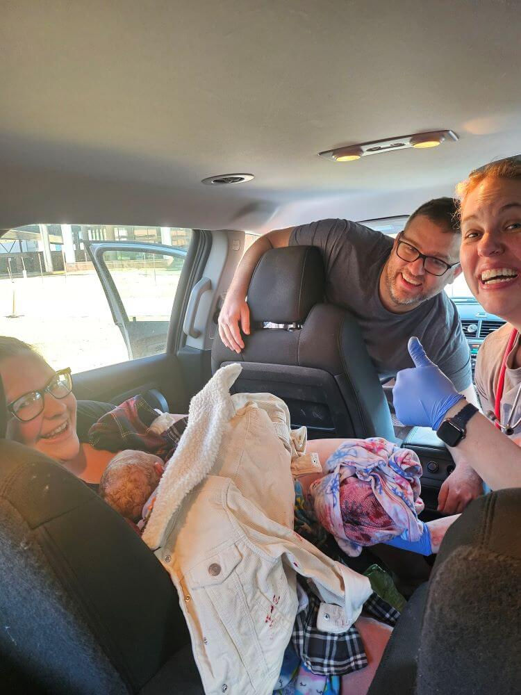 Three smiling adults after an accidental birth in a car. Mom is holding baby under random clothes, dad is in the driver's seat looking back, and the midwife is holding the placenta and giving a thumb's up