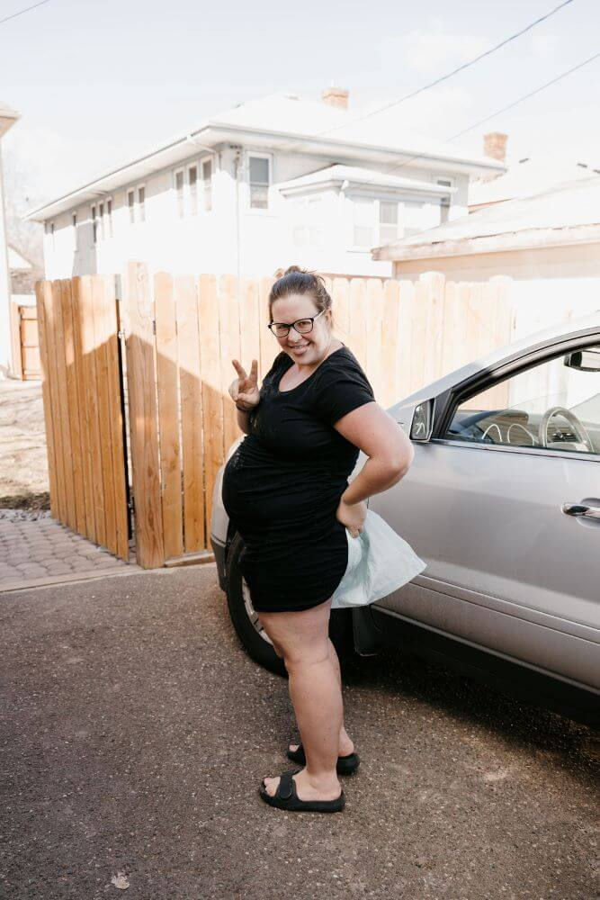 A mom stands outside after having just given birth in the car. She's wearing a black dress and holding a disposable pad between her legs. She's making a peace sign with her fingers and sticking out her tongue