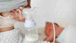 woman is pumping her breast and collecting milk in bags while a baby sleeps in the background