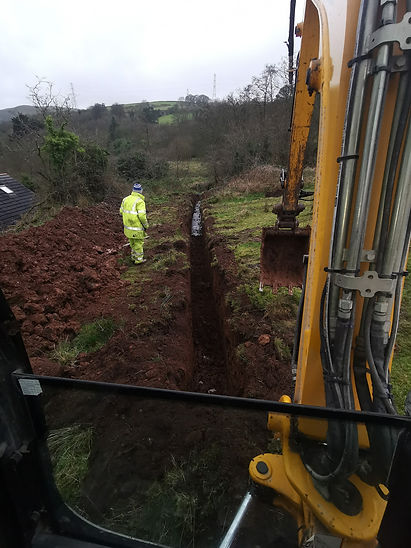 Worker in yellow suit and blue hat by a trench, excavator nearby