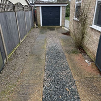 Narrow driveway with gravel path flanked by concrete, leading to a black garage doo