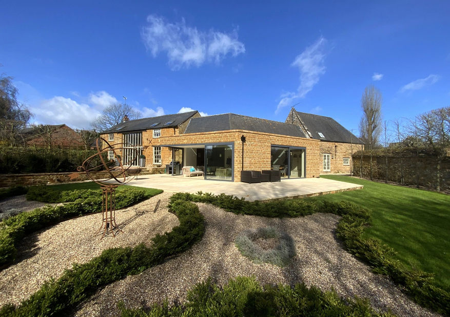 A modern stone house with large glass doors opens to a sunny patio and landscaped garden