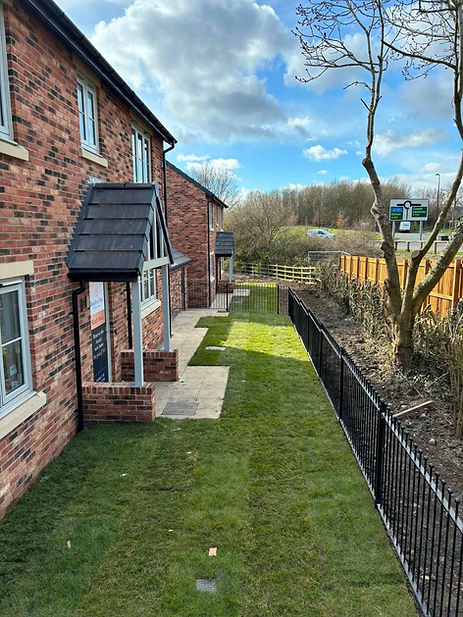 A newly turfed narrow garden path alongside a modern red brick house with black railings and a timber fence boundary.