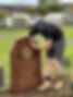 A man in work boots and a cap cleans a large, reddish gravestone in a cemetery
