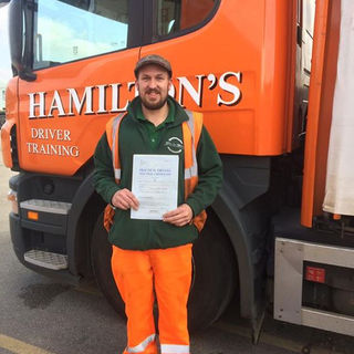 A man in an orange high-vis suit and green jacket stands in front of an orange Scania truck holding a pass certificate