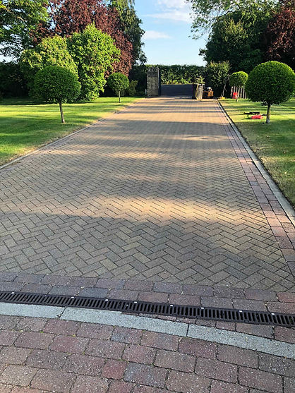 Long block paved driveway with red and grey borders leading to a house surrounded by green lawns