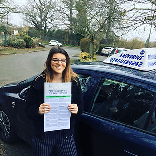 A smiling person stands outside, holding a Practical Driving Test Pass Certificate