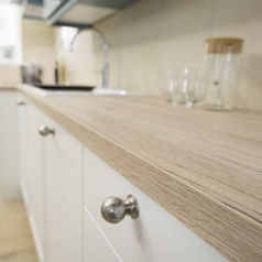 A close-up view of a light-colored wood grain countertop with round metallic knobs on white lower kitchen cabinets