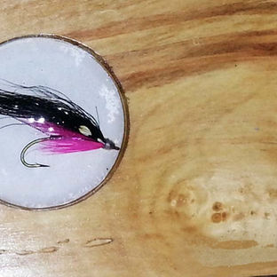 Close-up of a circular hand-painted illustration of a fishing fly embedded in natural polished wood grain