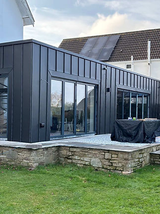 Modern black metal extension with large glass sliding doors, atop a stone patio