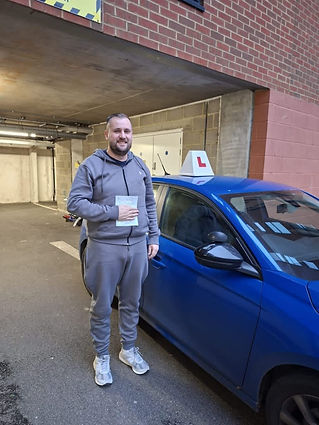 A smiling person stands beside a blue car with a learner's sign on top, holding a document