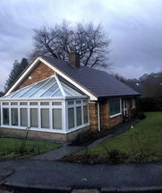Small brick house with large glass conservatory, lawn, and bare trees on a cloudy winter day