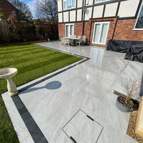 A modern backyard features a gleaming tiled patio, stone birdbath, and lush green lawn