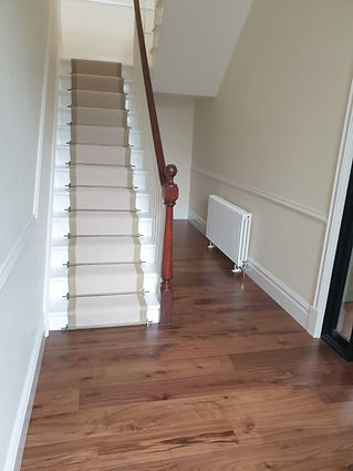 A wooden staircase with a beige carpet runner ascends in a narrow hallway with light-coloured walls