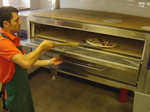 Pizza chef in an orange uniform carefully sliding a fresh pizza into a commercial deck oven