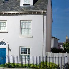 A white townhouse with a blue door, surrounded by shrubs and a black fence