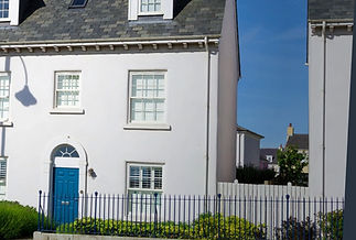 A white townhouse with a blue door, surrounded by shrubs and a black fence