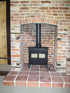 A black wood-burning stove sits on a raised, tiled hearth within a rustic red brick fireplace