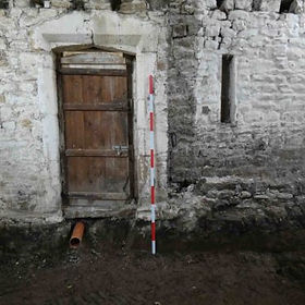 Old wooden door set in a rustic stone wall, with a vertical measuring rod beside it