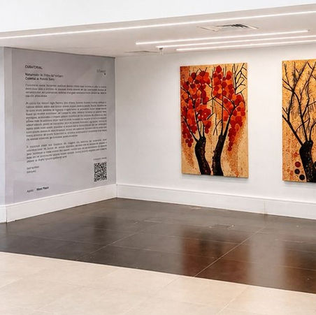 Parede branca exibe dois quadros de árvores com folhas vermelhas e fundo alaranjado. Placa com texto de exposição ao lado. Ambiente moderno.