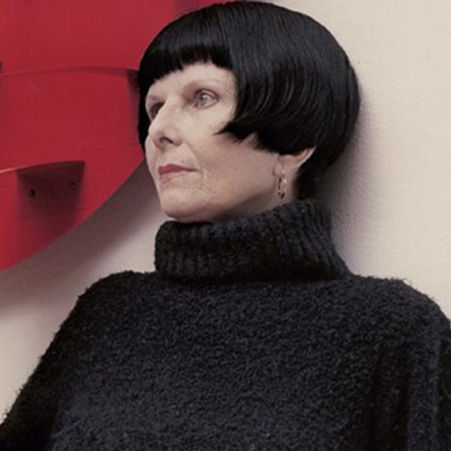 Retrato de Lygia Pape em ambiente expositivo, com corte de cabelo geométrico e roupa escura, diante de obra abstrata vermelha, imagem associada ao rigor formal e à pesquisa experimental da arte brasileira do século XX.