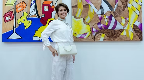 Mulher sorridente de branco posa em galeria diante de duas pinturas abstratas coloridas.