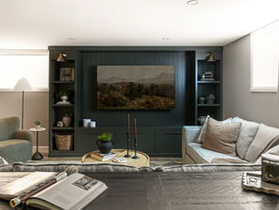 A cozy, modern living room featuring a built-in dark green entertainment center with open shelving and a mounted landscape painting above a hidden television. The shelves are decorated with books, plants, ceramics, and baskets. A neutral-toned sofa with textured pillows faces a round wooden coffee table styled with candles, a black vase, and stacked books. The space is softly lit with recessed ceiling lights and wall-mounted brass sconces.