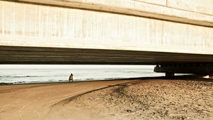 en eenzame figuur in de schaduw van een betonnen viaduct op een uitgestrekt strand in Muscat, Oman. Authentieke fotografie van Leon Bouwman voor 'Alone'. | A solitary figure in the shadow of a concrete viaduct on an expansive beach in Muscat, Oman. Authentic photography by Leon Bouwman for 'Alone'.