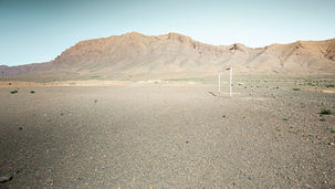 Een leeg, gebarsten sportveld tegen de achtergrond van het Atlasgebergte in Midelt, Marokko. Fine art fotografie van Leon Bouwman. | An empty, cracked sports ground against the backdrop of the Atlas Mountains in Midelt, Morocco. Fine art photography by Leon Bouwman.
