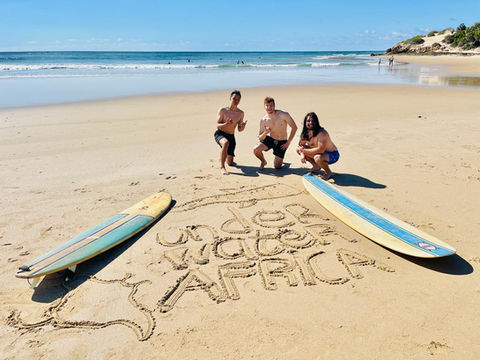 Three people kneel in the sand next to two surfboards framing the writing 'Underwater Africa' on the beach