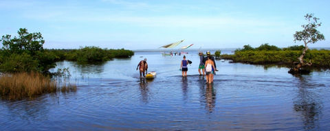 A group of people ushes heavily laden kayaks towards traditional sailboats in the background